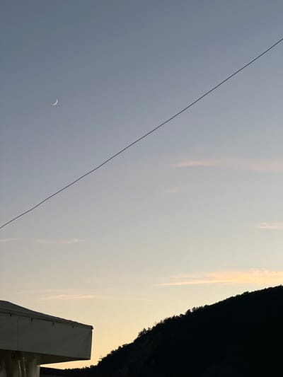 A crescent moon is visible in the dusky sky above Lermontovo, Russia, near a dark, tree-covered mountain range. A thin power line diagonally bisects the sky. In the foreground, the dark silhouette of a hillside is juxtaposed against a pale, yellowish sky. To the left, the side of a white structure, possibly a building or tent, is partially visible. The overall scene depicts a serene twilight atmosphere.