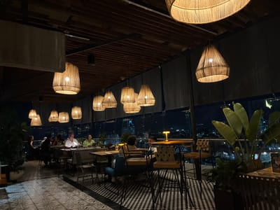 The image captures a nighttime scene within a high-floor or rooftop restaurant located in Floridablanca, Colombia, offering a panoramic view of the illuminated city skyline.
The establishment's interior features a dark, slatted wooden ceiling from which approximately fifteen to twenty woven, natural-fiber pendant lamps of various sizes are suspended, emitting warm, diffused light. The flooring consists of two distinct tile patterns: a lighter geometric pattern in the foreground transitioning to a black and white checkerboard pattern in the mid-ground.
Approximately 10-15 individuals, primarily adults, are visible within the dining area. They are seated at various tables and chairs, including standard dining sets and high-top tables with stools. Interactions include conversations and dining. A man wearing a light-colored shirt is seated at a central table, observed looking downward, possibly at a menu or mobile device. Another man in a green shirt is seated opposite him.
The environment is designed with large, potted green plants featuring broad leaves, positioned near the perimeter. The restaurant area is enclosed by glass partitions, offering unobstructed views of the city lights below. Dark roller blinds are partially deployed along these partitions. Additional localized illumination is provided by small, glowing table lamps positioned on some of the tables.