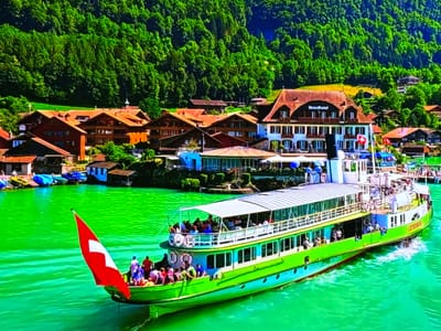 This media file depicts a scenic view of a passenger boat on a vibrant green body of water, likely a lake. The boat, painted in shades of lime green and white, is filled with people enjoying the ride. A Swiss flag is prominently displayed on the front of the boat, suggesting a European setting.

In the background, a picturesque village with traditional wooden houses and red-tiled roofs lines the shore. Behind the village, a dense, verdant forest covers a steep hillside, creating a dramatic backdrop. The architecture of the buildings, particularly the hotel with "Strandhotel" signage, points to a tourist destination.

The time of day appears to be sunny and fair, with clear skies contributing to the bright, almost tropical hue of the water. The presence of life rafts and the overall atmosphere suggest a leisurely recreational activity, perhaps a scenic tour or a ferry service. The movement of the boat creates small wakes on the water, indicating it is actively navigating. The people on board are casually dressed, enjoying the pleasant weather and the surrounding natural beauty.

The visible text on the boat, "LÖTSCHBERG", is likely the name of the vessel. The "Strandhotel" text on the building identifies it as a lakeside hotel. The Swiss flag is a clear indicator of the country. The scene evokes a sense of tranquility and vacation.
