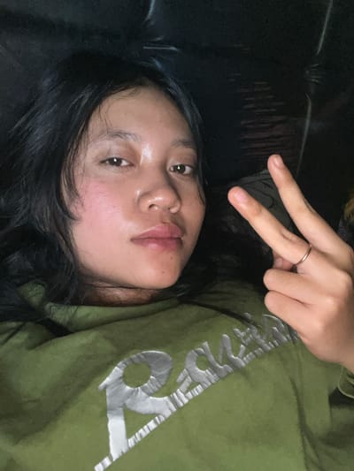 A young woman is shown in a close-up, selfie-style photograph, lying down and looking directly at the camera. Her long, dark hair frames her face, and she has fair skin with a slightly flushed complexion. She is wearing a green hooded sweatshirt with a metallic silver graphic design on the front. Her right hand is raised, with two fingers extended in a peace sign. A thin, gold ring is visible on her ring finger. The background is dark and indistinct, suggesting an indoor setting. The image is well-lit, focusing on the woman's face and upper body. There are no visible elements that place this image in Jambearum, Indonesia; the description is limited to the visual information present.