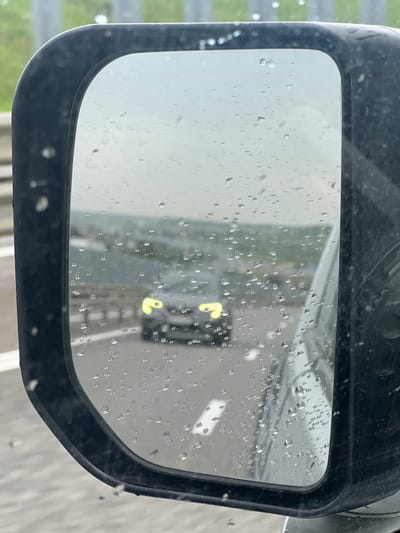 A rain-streaked side-view mirror of a vehicle reflects a fire engine driving behind. The mirror itself is black. Raindrops cover the surface of the mirror, obscuring the clarity of the reflection. The fire engine appears to have lights flashing. The environment outside the mirror shows a wet road surface and indistinct surroundings. The location is unknown.