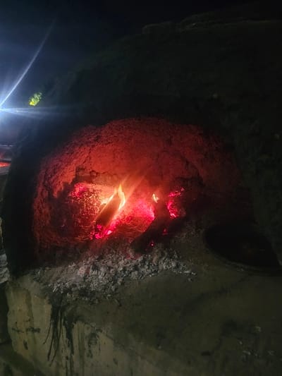 A close-up shot captures the interior of what appears to be a fireplace or furnace, the rough, textured walls partially illuminated by a reddish glow emanating from burning wood within. The firelight casts intense, uneven illumination, revealing charcoal deposits and soot-stained surfaces. A dark, circular opening is visible at the top. Lower in the image, what appears to be a flashlight beam is pointed toward the foreground.