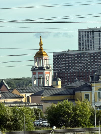 This image, captured in Kotelniki, Russia, depicts a street-level view dominated by urban architecture and lush greenery. The scene is characterized by several prominent buildings, including a tall, modern high-rise with a grid-like facade of windows and a neighboring building with a red and brown checkered pattern. Adjacent to these is a more traditional structure, a church bell tower with a golden dome, adorned with red and white detailing and arched openings. The foreground is crisscrossed by numerous dark wires, possibly power lines, which create a layered effect across the frame.
Behind the buildings, a dense canopy of green trees suggests a natural element within the urban landscape. The weather appears clear, with a bright sky, indicating it's likely daytime. Several cars are visible, parked along a street or road, hinting at typical urban activity. Streetlights are also present, one prominently positioned near the trees. No people are clearly discernible in the image. The overall impression is of a bustling city environment where modern and historical architecture coexist with natural elements. The orientation of the image is vertical.
