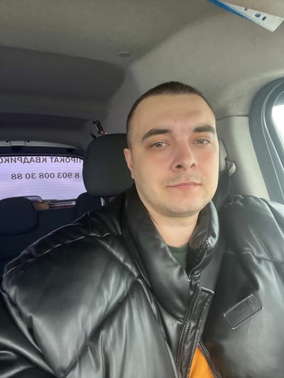 A male individual is positioned in the foreground of a vehicle interior in Rogachovo, Russia. He has short dark hair, light brown eyes, and a fair complexion, looking directly at the viewer with a neutral expression. He is wearing a black quilted puffer jacket over a dark green shirt, with a dark seatbelt visible across his right shoulder and chest. The vehicle's interior features a light gray headliner and a headrest behind the subject. Through the rear window, reversed Cyrillic text advertising "ПРОКАТ КВАРТИР" (Apartment Rental) and a contact number, "8 803 308 88", are clearly visible. The exterior environment, partially visible through the vehicle's right side window, is blurred and indistinct. The individual is stationary, appearing to be the sole occupant depicted.
