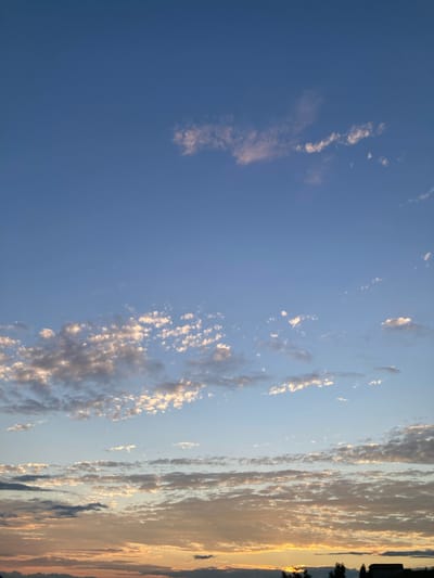 A sunset casts a warm glow over the sky, illuminating scattered clouds in a serene outdoor scene. The sky transitions from a deep blue at the top to a softer, lighter hue near the horizon, where streaks of orange and yellow paint a vibrant picture. The clouds, varying in size and density, are tinged with pink and gold, some appearing wispy and delicate, while others are more substantial and textured. In the distance, silhouetted against the colorful sky, are the outlines of trees and a building, adding a sense of place to the tranquil atmosphere. This image appears to be taken from Avanos İlçe Merkezi, Turkey, a location known for its picturesque landscapes and vibrant sunsets.