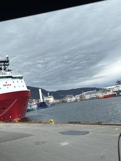 The image shows a view of the city of Bergen, Norway, from a dock. In the foreground, a large red ship with "SEA1" printed on its side is docked. To the right, a gray concrete dock is visible with some wet spots. To the left of the ship is the harbor with some buildings, a mountain, and some clouds in the background. In the harbor, another ship is visible.