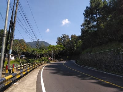 The image depicts an outdoor scene in the city of Hønefoss, Norway, on a clear, sunny day. The primary subject is an asphalt road curving from the lower center-left towards the mid-right. The road is marked with a solid white line on its left edge and a double solid yellow line, indicating a no-passing zone, near its center.

To the left of the road, a concrete sidewalk abuts a black and yellow striped metal guardrail. Further left, two utility poles, one light grey and one darker, stand prominently, supporting numerous overhead power lines or communication cables that stretch across the upper left quadrant. Beyond the guardrail, a cultivated area with visible soil and rows of low-lying vegetation is present. In the mid-distance on the left, a small white building with a blue section on its roof is partially visible, with what appears to be a white bus or large van parked nearby.

To the right of the road, a steep, heavily vegetated embankment rises, featuring a stone retaining wall at its base. Sections of this wall are topped with green netting or fencing. The embankment is covered in dense, dark green foliage, including tall evergreen trees on the upper right.

In the background, rolling hills or mountains covered in lush, dense green forest dominate the horizon under a bright blue sky with two distinct white cumulus clouds. No people or active vehicles are visible in the scene.