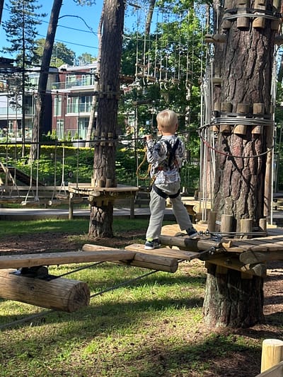 This media file captures a young boy navigating a rope course suspended between trees. The boy, wearing a light-colored shirt with a black and white pattern and grey pants, is safely harnessed and appears to be mid-challenge, with one foot on a wooden plank and the other extended forward. He has blond hair and is looking down, focused on his movement.
The rope course consists of natural trees as support, with wooden planks and ropes forming the obstacles. The scene is set outdoors in a park-like environment, with abundant greenery, including trees and grass, suggesting a daytime setting under clear skies. The sun casts shadows, indicating it's likely a sunny day. In the background, through the trees, a multi-story building with a reddish-brown facade and numerous windows is visible, providing a clue about the surrounding urban or suburban landscape.
The overall impression is one of outdoor recreation and adventure, with the boy engaged in a physical activity that requires balance and concentration. The presence of safety equipment like the harness emphasizes the controlled nature of the activity. The location context of Jūrmala, Latvia, suggests a coastal town known for its natural beauty and recreational opportunities. There is no visible text in the media file.