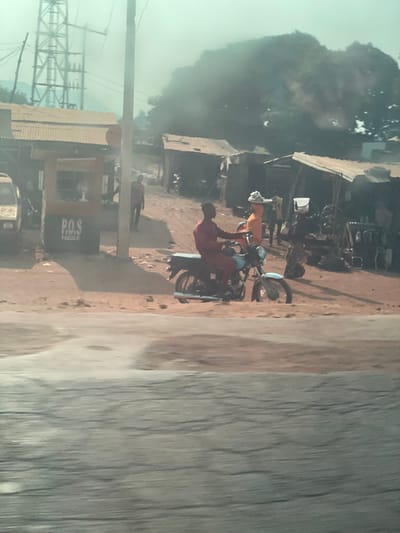 This image depicts a bustling roadside scene in Koton Karifi, Nigeria. The foreground shows a textured, grey paved road. Immediately beyond, a wide expanse of reddish-brown dirt serves as a common area for pedestrians and structures.
On the far left, a tall, metallic electricity transmission tower is visible against a hazy, light-green sky. Below it, an older, light-colored vehicle is partially parked near a standalone yellow kiosk marked "P.O.S" on its front. A vertical utility pole stands nearby.
In the midground, a man wearing a reddish-brown outfit is seated on a black motorcycle, facing right, on the dirt path. Slightly ahead of him, a woman in an orange top and dark skirt walks right, balancing a large, light-colored woven basket on her head. Further right, near a series of makeshift market stalls with corrugated metal roofs, another woman is partially visible, carrying a white bucket on her head while interacting near displayed goods. Multiple other blurred figures are present in the background, walking or standing among the stalls.
The background features a large, dark green tree, partially obscured by significant atmospheric haze or smoke. The overall environment is dusty, with strong sunlight casting distinct shadows from the people and structures onto the dirt ground.