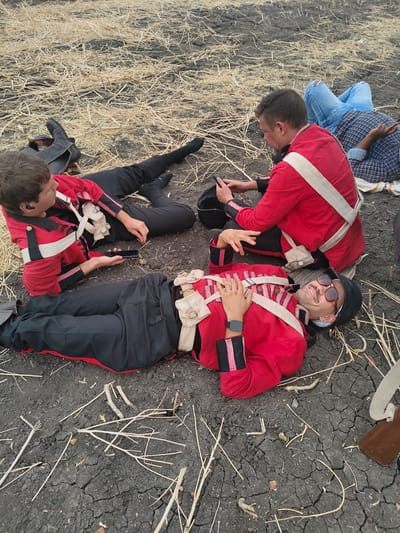 The scene depicts four individuals resting on dry, cracked, dark soil covered with scattered light-colored plant stubble in Kottalapalli, India. Three individuals are dressed in red military-style jackets with black cuffs and pink stripes, white cross belts, and black trousers, resembling historical uniforms. The individual in the immediate foreground lies supine, wearing a black cap, dark sunglasses, a mustache, and a smartwatch on his left wrist, smiling towards the viewer. His right hand rests on his chest. To his left, another male reclines, wearing a similar uniform and an earpiece, engrossed with a black smartphone. Behind him, a pair of black boots rests on the ground. To the center-right, a third male sits, also in the red uniform, looking down at a smartphone he holds. In the background, partially obscured, a fourth individual lies prone, wearing light blue ripped jeans and a blue and white checkered shirt. No active dynamic interactions are observed between the subjects; all appear to be at rest.