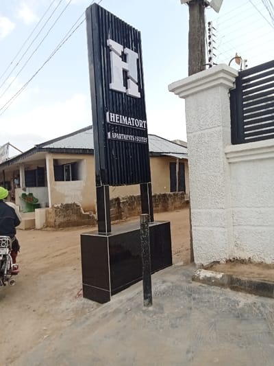 The image shows a sign for "Heimator Apartments/Suites" at 5.864 latitude and 5.701 longitude in Sapele, Nigeria. The sign is a black rectangular structure with a large white "H" on the top and the text "Heimator Apartments/Suites" in white lettering on the bottom. The sign is mounted on a black tile pedestal with a black metal pole in the middle. The sign is located in front of a white wall with a black gate to the right, and a one-story building to the left. There is a dirt road in front of the sign and a person on a motorcycle in the background. The sky is overcast. The sign is mounted on a concrete base with a black metal pole supporting the sign. The surrounding area appears to be residential.