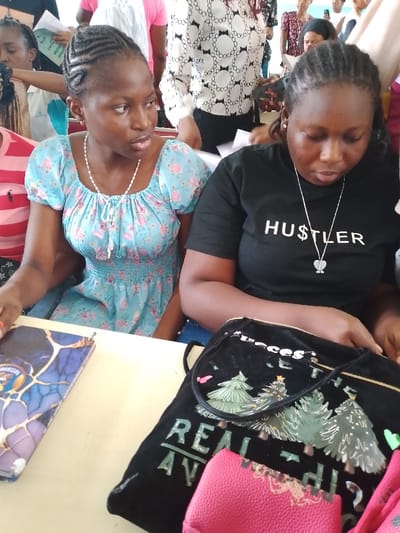 Two dark-skinned women are seated at a light-colored table. The woman on the left wears a light blue dress with pink flowers, and her hair is styled in cornrows. She is looking to the right. The woman on the right wears a black t-shirt with the word "HUSTLER" printed in white and a heart pendant necklace. She is looking down at something in her hands. A black bag with Christmas tree images and the words "Real" and "Success" in English sits on the table. A notebook with a blue cover sits to the left of the bag. Other people are standing in the background. The scene appears to be indoors. Based on the prompt, the location is Umueshi, Nigeria.