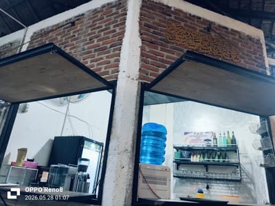 The image depicts the interior of a commercial establishment in Cempeudak, Indonesia. Two adjacent service windows are visible, each framed by a black metal structure with a wooden overhang. The dividing pillar is white and contrasts with the brick wall above. The wall features an Arabic inscription in gold. The left window reveals a coffee-making setup, including a small refrigerator, coffee grinder, and various containers. The right window contains a water dispenser with a large blue bottle, a shelf stocked with glasses, bottled beverages, and sachets. A badminton tournament poster is also affixed to the wall.