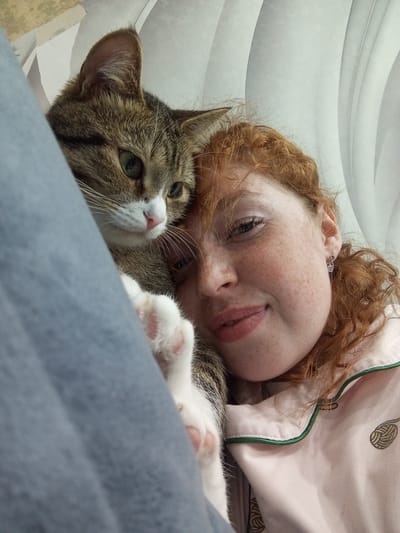 A light-skinned person with freckles and red hair is lying down wearing a light pink pajama top with a yarn ball design. A tabby cat with green eyes and white paws is on top of the person. A gray blanket is partially covering the upper left corner of the image. The background is white with curved lines. The location is not determinable from the visual elements of the image.