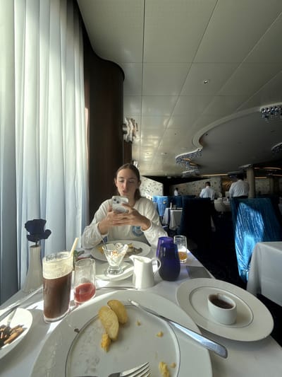 The image depicts a contemporary dining room scene in a restaurant located in the city of Rum Cay, Bahamas. In the foreground, a young Caucasian female with light brown hair is seated at a white-clothed table, facing slightly right. She wears a white long-sleeved shirt with a small emblem on the chest and is actively holding and looking at a white smartphone with both hands, her lips slightly pursed.
On the table in front of her, a partially consumed meal is visible. To her immediate right, a white plate contains a clear glass dish with a dessert-like item, next to a larger dark food item. Further to the left on her table, a dark beverage in a tall glass with a straw, and a smaller glass with a reddish drink are present. Two dark blue tumblers and a clear glass with an orange beverage are also on the table. A small white pitcher stands between the glasses. To the far left, a dark blue decorative object, possibly a rose, is in a white vase.
In the immediate foreground, a white plate shows remnants of a meal, including two potato wedges and scattered crumbs, with a fork and knife resting on it. To the right of this plate, a small white oval dish containing a dark sauce or condiment sits on another white plate. Further to the left, a plate with small, dark items is visible.
The restaurant's interior features bright natural light entering from large windows on the left, filtered by sheer white vertical curtains. Dark brown structural pillars or wall panels are present. The ceiling is white and perforated, with recessed lighting. Architectural elements further into the room include curved white structures with ornate blue crystal or glass light fixtures hanging from them. In the background, multiple tables covered with white tablecloths are set with dark blue upholstered chairs. Several individuals, likely restaurant staff wearing white shirts, are visible moving or standing in the background. Other patrons are also seated further back in the dining area. The overall ambiance suggests an upscale dining establishment.