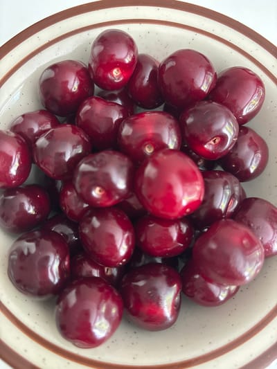 A close-up, overhead shot shows a bowl of dark red cherries. The cherries are plump and glossy, with some reflecting light and others appearing slightly darker. They are piled high in a shallow, ceramic bowl with a pattern of thin brown and beige stripes along the rim. The background is a plain, light color, possibly a countertop or table. The scene appears to be indoors, in a domestic setting. There are no people visible in the image. The location in Лунненскі сельскі Савет, Belarus, is not discernible from the image itself, as it contains no geographical markers.