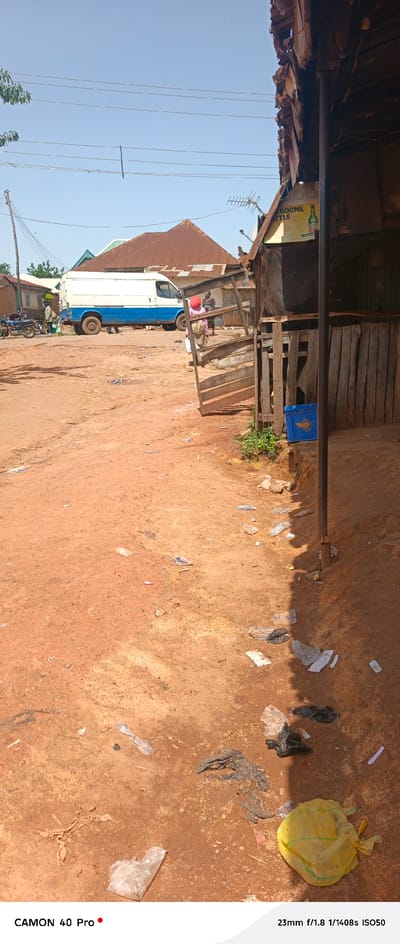 The image depicts an outdoor scene in Barikin Gidar Kanat, Nigeria. The foreground is dominated by a dirt road covered in debris, including plastic bags and other refuse. To the right, a rustic wooden stall with a corrugated metal roof is visible, with a blue plastic crate on the ground in front of it. Further into the scene, a white van with a blue stripe is parked on the dirt road. Several people are present, with two individuals standing near the van and another person, possibly a woman with a red head covering, standing to the right of the van. In the background, various buildings with corrugated metal roofs are scattered across the landscape, under a clear blue sky. Power lines are strung across the upper portion of the image. The overall impression is one of a dusty, unpaved area with signs of commercial activity and general litter.