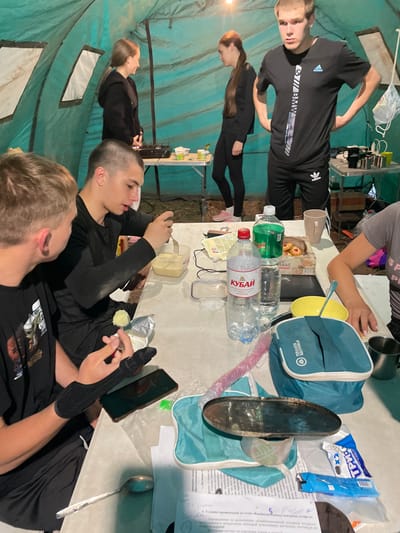A group of four young adults are gathered inside a teal tent, suggesting an outdoor or camping scenario in Nikolayevka, Russia. A young man in the foreground on the left is looking away from the camera, with his left arm bandaged. He is seated at a table with a phone and other items. To his right, another young man is eating from a plastic container with a fork, also seated at the table. In the background, a young woman in a black hoodie stands near a portable stove, and a young man in a black t-shirt and pants stands facing the camera, his hands on his hips. The table is laden with food items, water bottles, and various containers, indicating a meal or snack is in progress. The tent's interior is dimly lit, with light emanating from above and through window openings in the tent fabric.