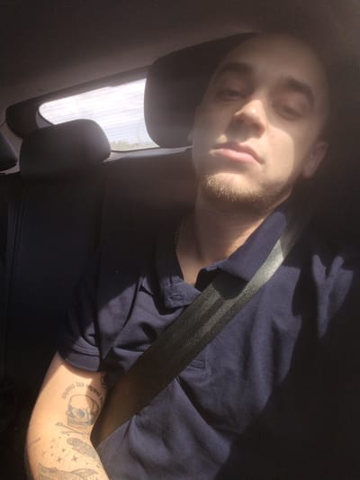 In Tolokonnikovo, Russia, a bald man with a goatee is seen from the chest up, sitting in the back seat of a vehicle. He is wearing a dark blue polo shirt and a seatbelt is across his chest. Sunlight streams in from the right, illuminating the left side of his face, creating harsh shadows. His left arm, visible on the left side of the frame, is adorned with tattoos, including a skull with text above it, and other designs. The background consists of dark car upholstery and a glimpse of the sky through the rear window.