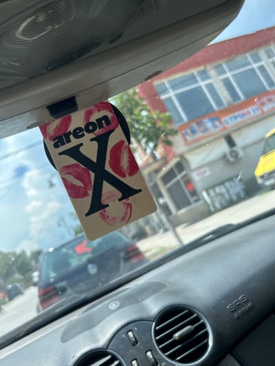 The image, taken from inside a car, shows a hanging air freshener with the brand name "areon" and a large "X" mark, adorned with red lipstick kiss prints. The air freshener is suspended from the car's rearview mirror assembly. Outside the car, a blurry cityscape is visible, including a building with a red facade and large windows, and another yellow vehicle. The dashboard of the car is partially visible, featuring an air vent and the "SRS AIRBAG" indicator. Based on context from other images in this set, this scene is located in the city of Krivodol, Bulgaria.