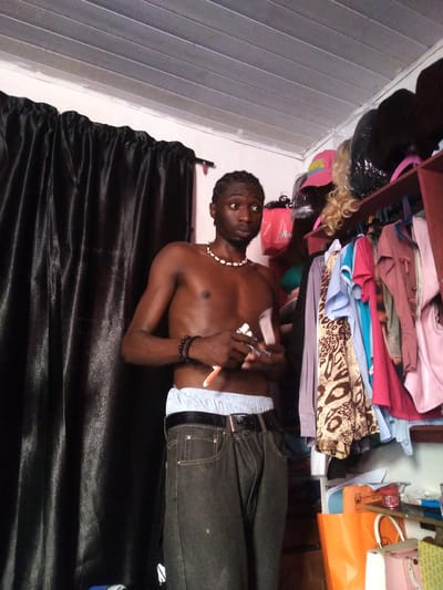 A young Black male stands shirtless in a cluttered room with a black curtain to his left and shelves full of clothes and wigs to his right. He wears dark gray pants and light blue underwear. He has a beaded necklace and two beaded bracelets on his left arm. He is holding two small, white objects in his right hand, possibly a lighter and a small pill bottle. The room appears to be a storage area or a dressing room, with clothes hanging on hangers and stacked on shelves. The ceiling is made of corrugated metal. The image was taken in Kassa Market, Nigeria.