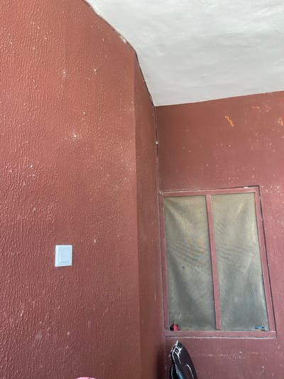 A low-angle shot reveals a section of a building interior, painted in a textured, reddish-brown color. A rectangular window with a screen is set into the wall. The screen is divided by a horizontal bar. At the bottom of the image, a white electrical outlet cover is visible on the floor. The floor has the same textured reddish-brown finish as the walls. A section of the adjacent wall is painted white. Part of a black object is visible in the upper right of the image. The location is Sabo, Nigeria.