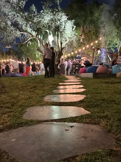 This media file depicts an outdoor evening gathering in Nir Eliyahu, Israel. The scene is illuminated by numerous warm string lights strung overhead and spotlights highlighting the foliage of large trees, creating a festive and inviting ambiance under a dark night sky.
In the foreground, a stone path consisting of irregularly shaped stepping stones extends diagonally across a lush, deep green grassy area. The path reflects the warm light, and fine moss or grass grows between the stones.
To the left, large trees with gnarled branches and dense green foliage are prominent, some with white, antler-like decorations hanging from their branches. These trees are lit from below, accentuating their forms against the dark sky. Other denser trees are visible in the background.
Numerous people are present, socializing and observing the event. Many individuals are standing and mingling, while others are seated on colorful beanbags or cushions (pink, blue, red, purple) arranged on the grass. Their attire appears casual. In the mid-ground, a group of people are seen standing and looking towards a more brightly lit area in the background.
In the background, a raised structure, possibly a stage, performance area, or bar, is visible with additional lighting. A faint haze or smoke is discernible around this area, suggesting an activity or performance is taking place there.
The overall atmosphere is relaxed and social, characteristic of an outdoor event or party. The time of day is clearly evening or night, indicated by the artificial lighting and the dark sky. No visible text is discernible in the image.