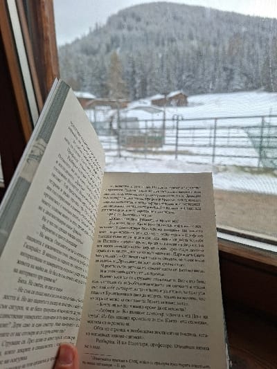A human hand, visible in the lower right, holds an open book displaying Cyrillic text on its two visible pages. The book is positioned in front of a window, with a dark wooden frame partially visible along the bottom and right edges. Beyond the window, a snow-covered landscape extends into the distance. A metal fence traverses the snowy midground, where two small, rustic wooden structures with reddish-brown roofs are present. In the background, a large mountain, densely covered with snow-laden evergreen trees, rises against an overcast sky. The scene is consistent with a winter alpine environment in Davos, Switzerland.
