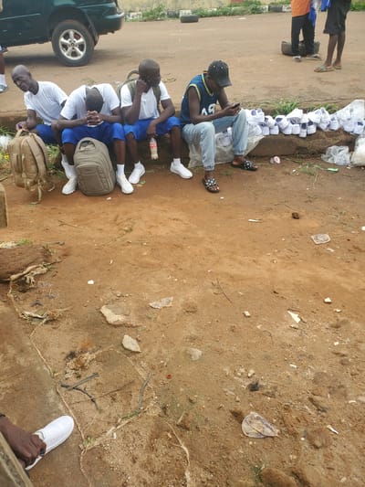 A group of young men, likely students, are seated on a dirt embankment next to a road. They are all wearing white t-shirts and blue shorts, with white socks and white sneakers. Three of the men have backpacks. One man, wearing a blue tank top and a black cap, is looking at his phone. In front of the seated men are piles of white shoes, some in plastic bags. The ground is dirt and sparsely covered with dry grass and debris. A dark green car is parked on the road in the background, and other individuals are visible further down the road. The scene appears to be outdoors, likely in a rural or semi-urban area within Bahit, Nigeria.