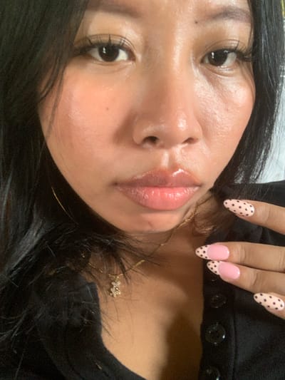 The image shows a close-up of a person's face and hands. The person has dark hair and is wearing makeup, including mascara and lip gloss. Their nails are painted pink with black polka dots. They are wearing a black shirt. The background is blurred. No specific location in Jajag, Indonesia, can be determined from the image.