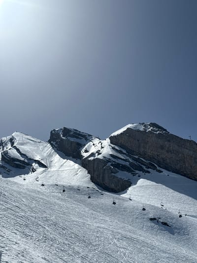 The image depicts a mountainside in La Giettaz, France, featuring a steep, snow-covered slope with visible ski tracks. A rocky cliff face is partially covered in snow and ice. A series of ski lift chairs ascend the slope, suspended by cables. The sky is a pale blue, with a bright light source visible in the bottom left corner.