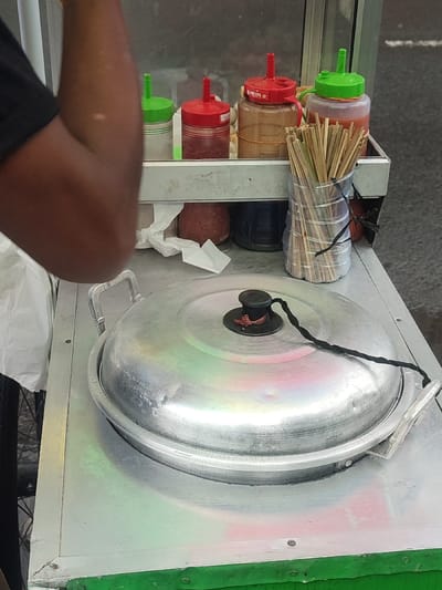 The image shows a food stall in Kecamatan Bangil, Indonesia. The stall is a small cart with a metal top. A large, round, silver pot with a lid sits on top of the cart. The lid is closed, secured with a black cord. The cord is tied to a knob on top of the lid. A person is standing behind the cart, their arm resting on the metal top. There are several bottles of condiments on a shelf above the pot. The bottles are filled with red and white liquids, as well as a brown liquid, likely sauces. A glass jar containing a bundle of wooden skewers is next to the bottles. The stall is located on a paved street. A person's foot can be seen at the bottom of the image, suggesting that the person is standing behind the stall, perhaps working.