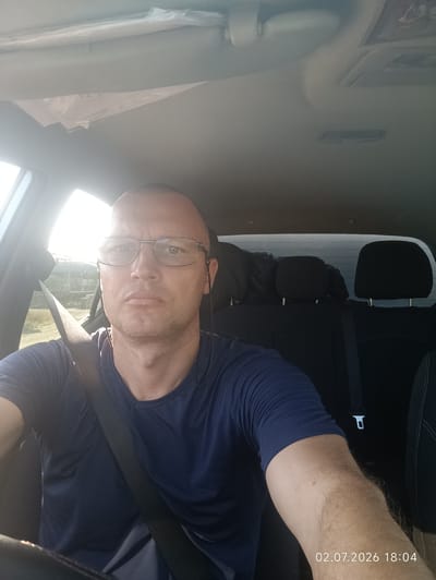 A selfie taken from the driver's seat of a car shows a bald, middle-aged man with glasses looking directly at the camera. He is wearing a dark blue V-neck t-shirt and a black seatbelt. The car's interior is visible, including the dashboard, steering wheel, and the backseats with headrests. Sunlight streams in from the left side of the car, creating a bright glare. The scene appears to be an outdoor setting with greenery visible through the car window. A timestamp in the bottom right corner of the image reads "02.07.2026 18:04".
