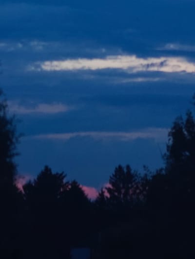 The image depicts a nighttime or twilight landscape with a dark sky filled with clouds. The sky is predominantly a deep blue, with lighter streaks of white and pink clouds visible. Silhouetted trees form a dark line across the lower portion of the image, obscuring the ground. There are no people, animals, or specific structures clearly identifiable in the scene. The overall impression is one of a natural environment at dusk or dawn. The location in Hadiach, Ukraine, is not discernable from the visual information provided in the image.