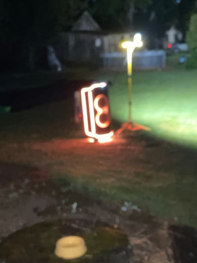 In the city of Maiņas, Latvia, an outdoor scene unfolds at night. The focal point is a large, cylindrical object adorned with illuminated, orange neon-like numbers "3" and "8." This object appears to be a speaker or some form of portable entertainment device. To the right of the object, a bright, yellow light emanates from a tall pole supported by a tripod stand. The ground is covered in grass, dimly lit by the surrounding lights and a distant, out-of-focus light source. In the background, a blurred structure resembling a building with a triangular roof is visible, along with hints of a fence and what might be a swimming pool. The overall atmosphere is dark and somewhat blurry, suggesting the image was captured in low-light conditions.