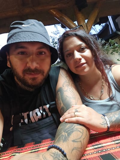 The media file is a close-up, high-angle selfie featuring two people, a man and a woman. The man is positioned in the upper half of the frame, his face angled towards the camera. He has a beard and is wearing a black hooded jacket and a dark bucket hat with a white logo on the side. The woman is in the lower half of the frame, her face also tilted towards the camera. She has long, dark hair and is wearing a grey tank top and a gold necklace.

Both individuals appear to be outdoors. The background shows natural elements like trees and branches, as well as what looks like a wooden structure, possibly a pergola or roof. The lighting suggests it is daytime, with soft natural light illuminating their faces.

Notable details include tattoos visible on both individuals' arms, suggesting a personal style. The woman is wearing rings on her left hand and a bracelet with white letter beads that spell out "INVI." The man has a dark beaded bracelet on his left wrist.

The overall mood of the image is casual and intimate, as if the couple is posing for a photo together. There is no clear indication of a specific event, but the setting suggests a relaxed outdoor gathering. Visible text includes the white logo on the man's hat and the letters on the woman's bracelet. The image appears to be taken in Dimitrovgrad Municipality, Serbia.