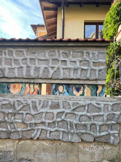 The image shows a section of a wall constructed with rough, grey stones and concrete. In the center of the wall, there is a vertical strip of colorful decorative tiles featuring abstract patterns and stylized human figures. The tiles display a palette of blues, greens, yellows, oranges, and pinks. To the left of the stone wall, there is a portion of a building with a yellow facade and a wooden-shingled roof. A window with a dark frame is visible on the yellow wall, reflecting a blue sky with some clouds. To the right of the stone wall, a rough concrete surface is visible. The scene appears to be outdoors, with green foliage at the top of the image, suggesting a daytime setting. The overall impression is of a rustic and artistic architectural detail.