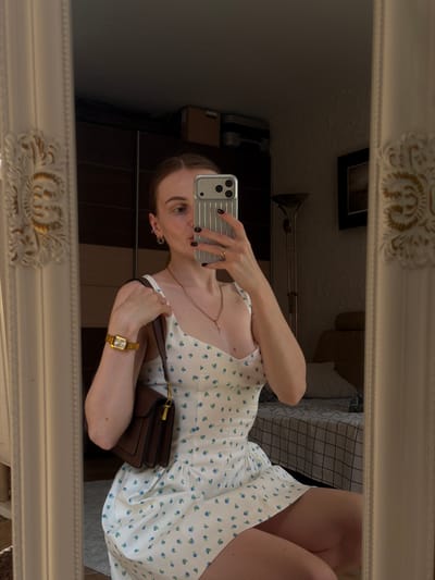 The image shows a woman taking a mirror selfie in what appears to be a bedroom. She is wearing a white floral print dress and holding a brown handbag. She is also wearing a gold watch and a necklace. The room is decorated with a large ornate mirror, a wardrobe, and a bed. There is also a floor lamp and a framed picture on the wall. The lighting is dim, suggesting it might be evening or the blinds are drawn. The woman's face is partially visible, and she is looking at her phone. The overall impression is one of a casual, domestic setting. The specific location in Gorzow, Poland, cannot be determined from the visual information provided in the image.