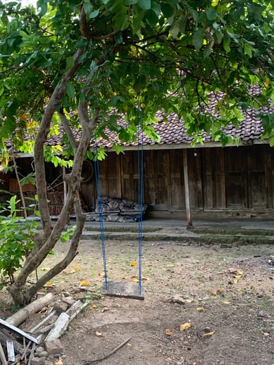 A wooden swing hangs from a tree with green leaves in front of a rustic wooden house with a tiled roof. The ground is covered with dirt, pebbles, and fallen leaves. Various discarded items, including bamboo poles, bricks, and metal pipes, are scattered near the tree. Cushions with a floral pattern are placed on the ground in front of the house. The scene appears to be an outdoor area in a rural or semi-rural setting, possibly within the city of Tanjungsepreh, Indonesia.