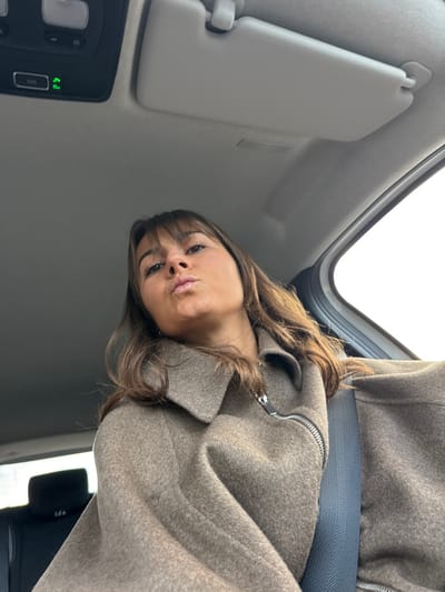 A woman with brown hair and bangs poses in a car. She wears a tan, zipped coat and a seatbelt. The car's interior features a sun visor, overhead console with buttons and lights, and a sunroof. The perspective is from below, looking up at the woman. The sky appears through the sunroof.