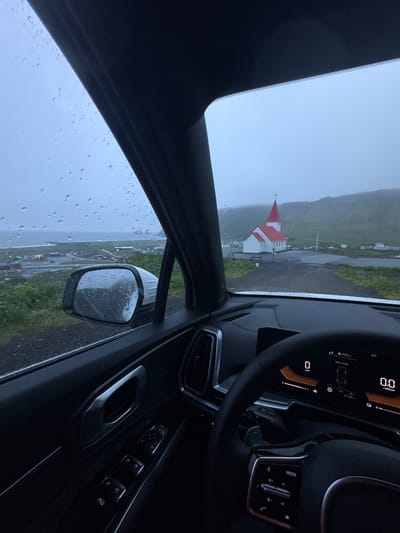 The image, taken from inside a car, shows a view of the Vikurkirkja Church in Vik, Iceland. Raindrops are visible on the car's windshield and side mirror. The church, with its characteristic red roof, stands prominently in the landscape, surrounded by green fields and houses. The interior of the car is visible, including the dashboard, steering wheel, and door panel. The car's dashboard display shows various readings, including mileage and engine speed. The overall atmosphere suggests a rainy and overcast day in Vik, Iceland.