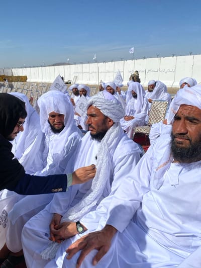 An outdoor scene in Mandisar, Afghanistan, depicts a gathering of numerous men under a clear blue sky. Most men are dressed in traditional white garments and head coverings, seated in rows. In the immediate foreground, a young man wearing a dark blazer over a light green shirt and a dark hooded top is leaning towards a seated man in white attire and a white turban. The young man's hands are engaged near the chest area of the seated man, possibly attaching or adjusting a small dark object, which appears to be a pin or badge, to his white garment. The seated man wears a dark watch on his left wrist and has a dark beard. To his right, another man with a full dark beard and white attire is seated, observing. In the mid-ground, rows of empty white plastic chairs are visible. The background features a tall, light-colored concrete or HESCO barrier wall topped with barbed wire. Several white flags with black Arabic script are visible on or behind the wall. Arabic script, partially legible as "الاسلامي امارات افغانستان" (Islamic Emirate of Afghanistan), is painted on the wall. A figure in camouflage attire is visible near the wall. In the distance, hills or mountains are discernible beneath the horizon.