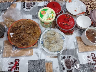 The image depicts a table set with a meal in Onaldy, Turkmenistan. The table is covered with a patterned tablecloth. On the table, there is a large dish of what appears to be stewed meat, a plate of dumplings or ravioli covered in plastic wrap, and a large tub of mayonnaise. Two red lidded pots are also present, along with several plates and bowls, some of which are empty or contain remnants of food. A cup of coffee with a heart-shaped foam design and coffee beans is visible on a saucer. The overall scene suggests a home-cooked meal.