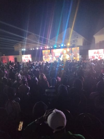 The image depicts an outdoor nighttime event taking place in Fagge D2, Nigeria. A large audience, consisting of hundreds of individuals, is gathered in an open area, predominantly seated in rows of chairs, facing a brightly lit stage structure.
The stage features a metal truss framework supporting multiple large LED screens. The central screens display a consistent image of a bearded male figure wearing a cap or head covering, holding a microphone, suggesting a performance or speech. Stage lighting fixtures are mounted on the truss, emitting visible beams of colored light (blue, green, purple, yellow, white) that extend upwards into the dark night sky. Additional screens are visible to the left and right of the main stage.
In the foreground, the backs of audience members are visible, mostly silhouetted against the stage lights. One individual in the immediate foreground wears a white baseball cap. Another person holds an illuminated mobile phone. Behind the stage, parts of buildings with light-colored roofs are discernable. The crowd extends into the deep background, indicating a substantial attendance.