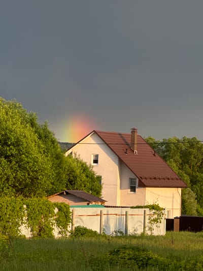 The image shows a house in Vysokinichi, Russia, with a partial rainbow visible on the left side of the building against a dark, overcast sky. The house is two stories with a brown roof, a chimney, and white siding. A white fence with vines growing on it runs along the right side of the house. Lush green foliage surrounds the house, partially obscuring it from view. A small outbuilding with a dark roof is situated next to the house. The foreground includes grass and more dense foliage.