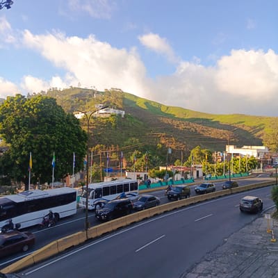 The image depicts a view of a multi-lane road in Carrizal, Venezuela, with a backdrop of a green, hilly landscape dotted with houses and trees. The sky is bright blue with scattered white clouds, suggesting it is daytime.
In the foreground, several vehicles are present on the road. A white bus with black-tinted windows is in the left lane, and another white bus with blue and yellow detailing is in the adjacent lane. A red motorcycle with two riders is visible between these buses. Further down the road, a dark-colored SUV is driving in the right lane, followed by a silver sedan and another dark-colored SUV. In the far right lane, a light-colored SUV is driving away from the camera. Yellow concrete barriers separate the road lanes from the pedestrian walkway and the embankment.
On the left side of the road, a large, mature green tree stands prominently. Several flags on poles are visible near the tree. Behind the tree and the road, buildings and more trees are situated on the lower slopes of the hills. The hills themselves are covered in green vegetation, with some areas appearing as terraced fields. Houses with white walls and red roofs are scattered across the hillside, with some appearing modern and others more traditional. A lamppost with multiple light fixtures is positioned on the left side of the image, near the edge of the road.
On the right side of the image, a yellow sign with red and white stripes stands near the edge of the road, possibly indicating a no-parking or no-stopping zone. Further back, more buildings and vegetation are visible. The overall scene suggests an urban environment integrated with a natural, hilly landscape.