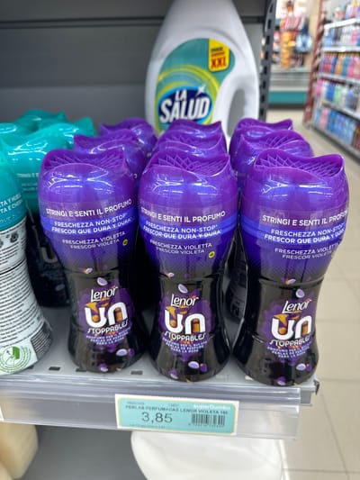 The image shows a shelf in a supermarket, likely in Murcia, Spain, displaying a variety of laundry scent boosters. In the foreground, several bottles of Lenor Unstoppables in the "Violeta" scent are prominently featured. These bottles are purple and black, with a distinctive ridged purple cap and a label that reads "Lenor" and "Unstoppables" in large white lettering. The labels also contain text in Italian and Spanish, such as "Stringi e Senti il Profumo" and "Freschezza Non-Stop." Some of the bottles have small purple flower petal graphics on them. Behind these, partially visible, are bottles of other laundry products, including teal-colored ones to the left and a large white bottle with a green and blue label featuring the "La Salud" brand and an "XXL" size indicator. The overall environment appears to be a retail setting with shelves stocked with consumer goods. A price tag is visible at the bottom of the image, indicating "PERLAS PERFUMADAS LENOR VIOLETA 195" and a price of 3,85.
