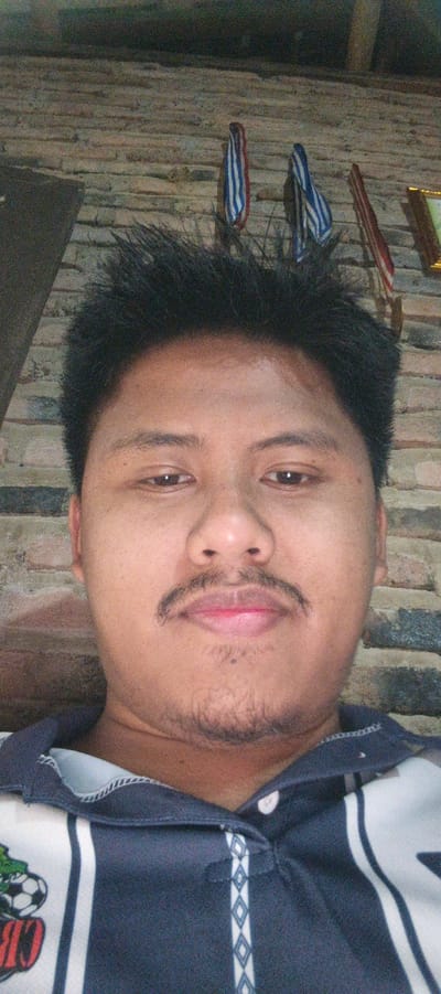 The image is a close-up selfie of a young man with dark, spiky hair, a mustache, and light stubble on his chin. He has brown eyes and is wearing a dark blue polo shirt with white stripes and a diamond pattern. Behind him, a brick wall is visible, with two small, colorful decorative items hanging on it, one appearing to be a red, white, and blue ribbon. The lighting suggests an indoor setting. The man's expression is neutral. The location is identified as Kemuning, Indonesia.