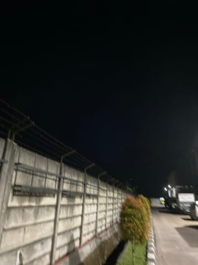 A nighttime outdoor shot shows a concrete wall topped with a barbed wire fence running horizontally across the lower half of the frame. To the right of the wall, a small, colorful bush stands near a paved walkway. In the background, streetlights illuminate a road and a parked truck. The sky above is dark. The scene is located in Gambut Barat, Indonesia.