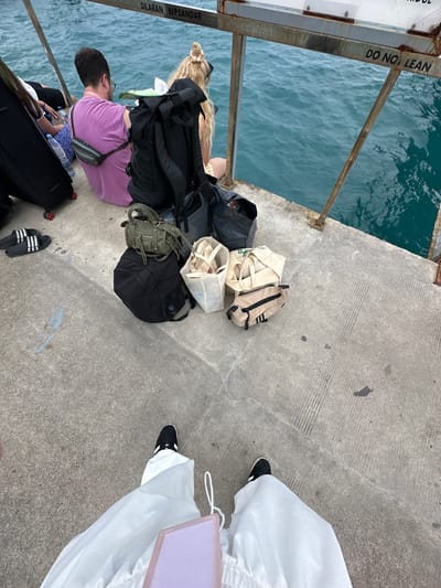 The scene is on a concrete pier in Kampung Toyapakeh, Indonesia, beside a body of water. There are several pieces of luggage and bags, including black backpacks, canvas tote bags, and a rolling suitcase. Two people are visible: one wearing a purple shirt and the other wearing white pants and black shoes. A sign with the phrase "DO NOT LEAN" can be seen. The environment suggests a travel or waiting situation.